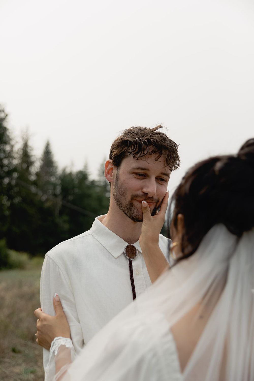 Groom smiling as bride gently wipes lipstick from his face during outdoor wedding portrait in forest setting.