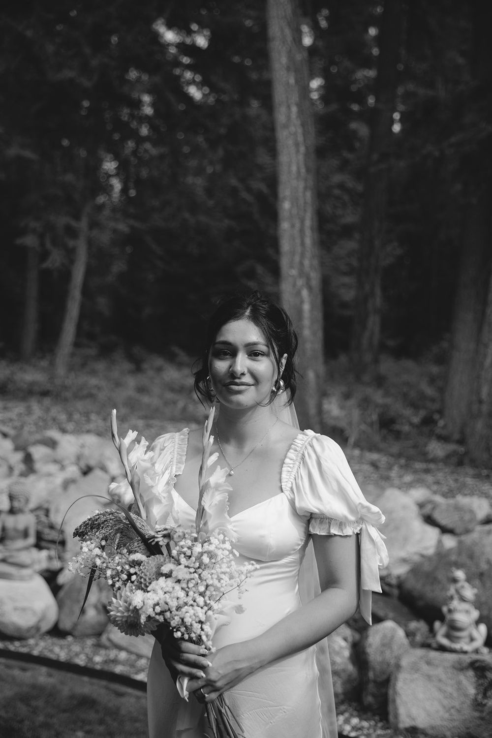 Black and white portrait of bride in white gown holding wildflower bouquet near stone garden.