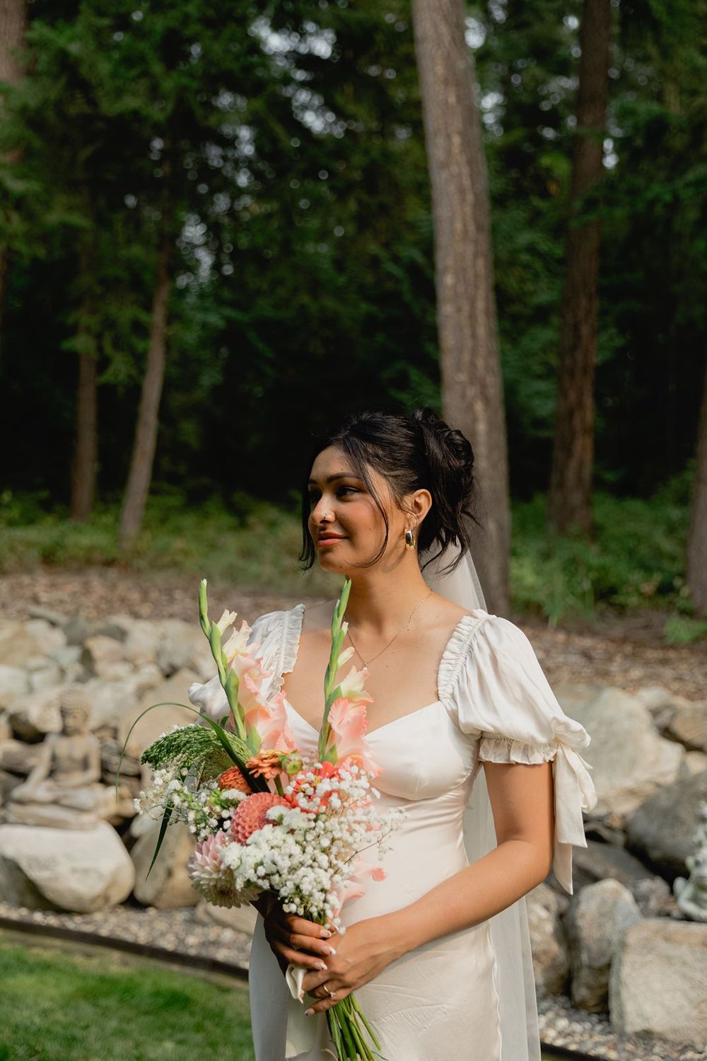 Bride in white puff sleeve gown holding pink and white floral bouquet in lush forest setting.