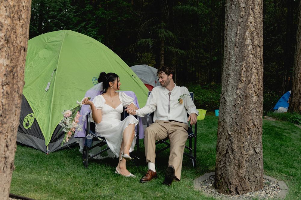 Bride and groom relaxing in camp chairs beside green tent in wooded campsite after wedding.