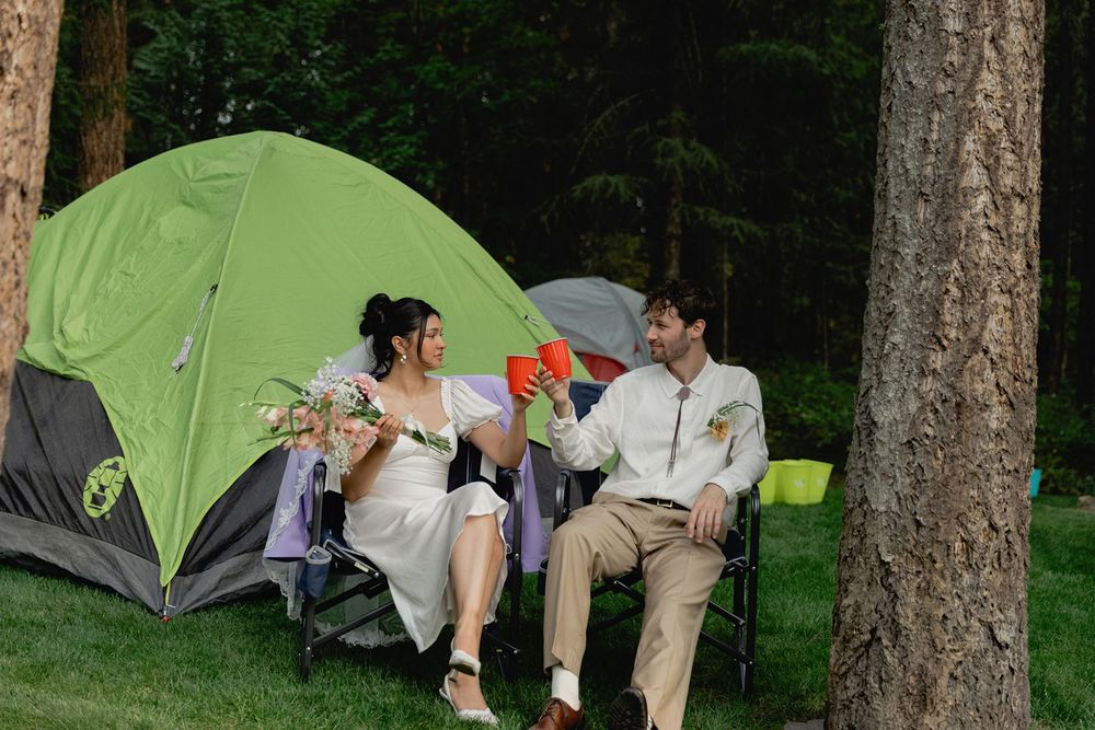 Bride and groom relaxing in camp chairs beside a green tent at their outdoor forest campsite wedding.