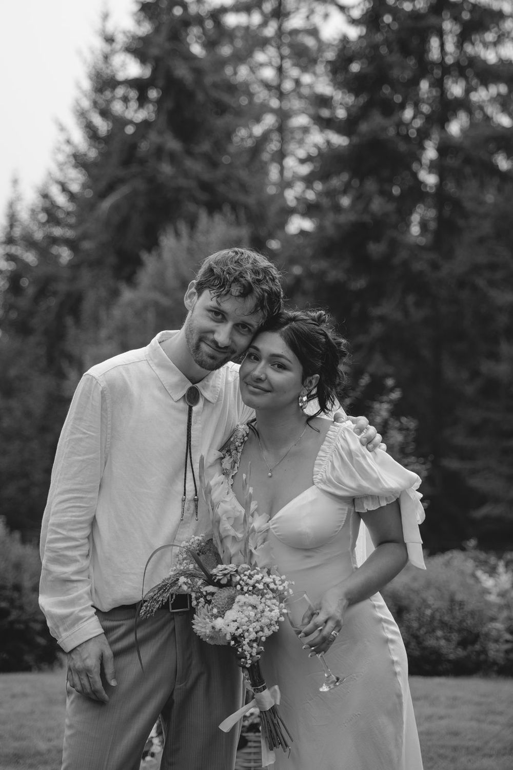 Black and white portrait of smiling bride and groom posing together outdoors with wildflower bouquet.
