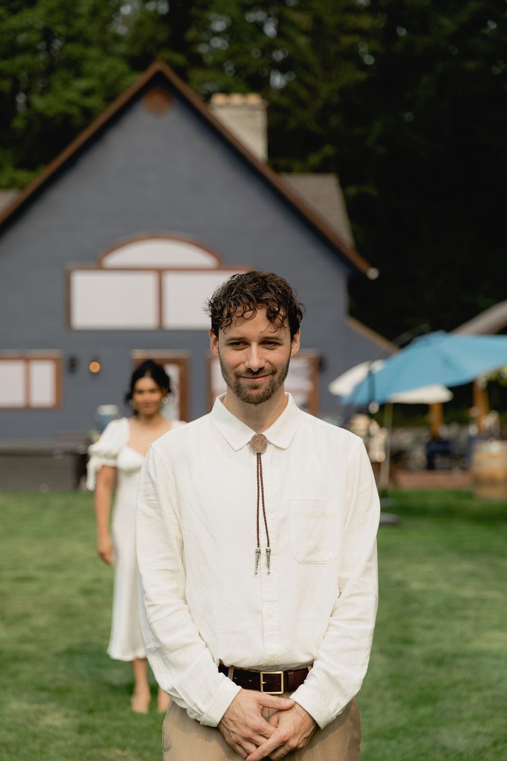 Smiling groom in white shirt with bolo tie standing outdoors at a blue estate venue.