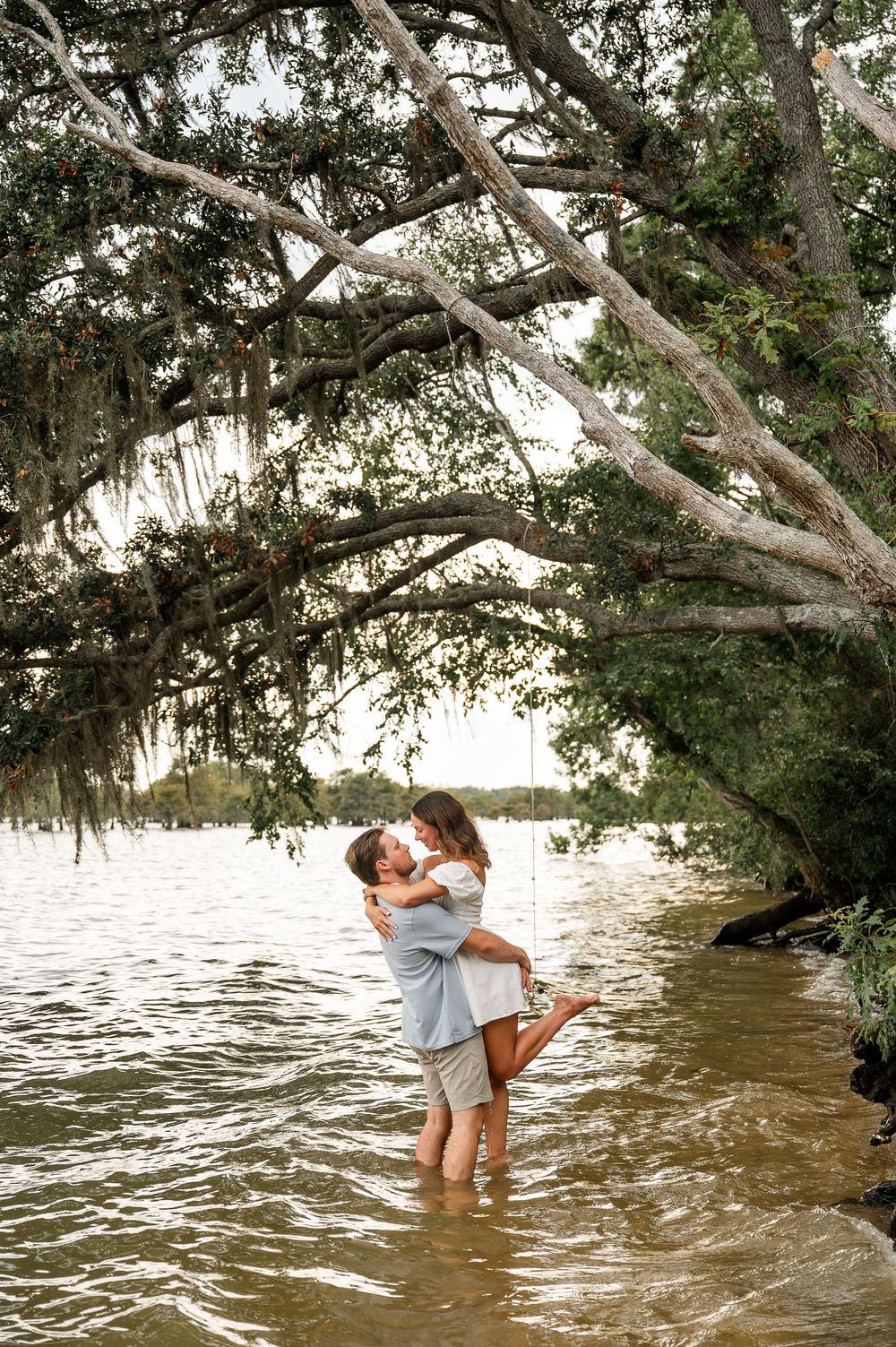 A couple shares romantic moments while standing in shallow water under a large oak tree.