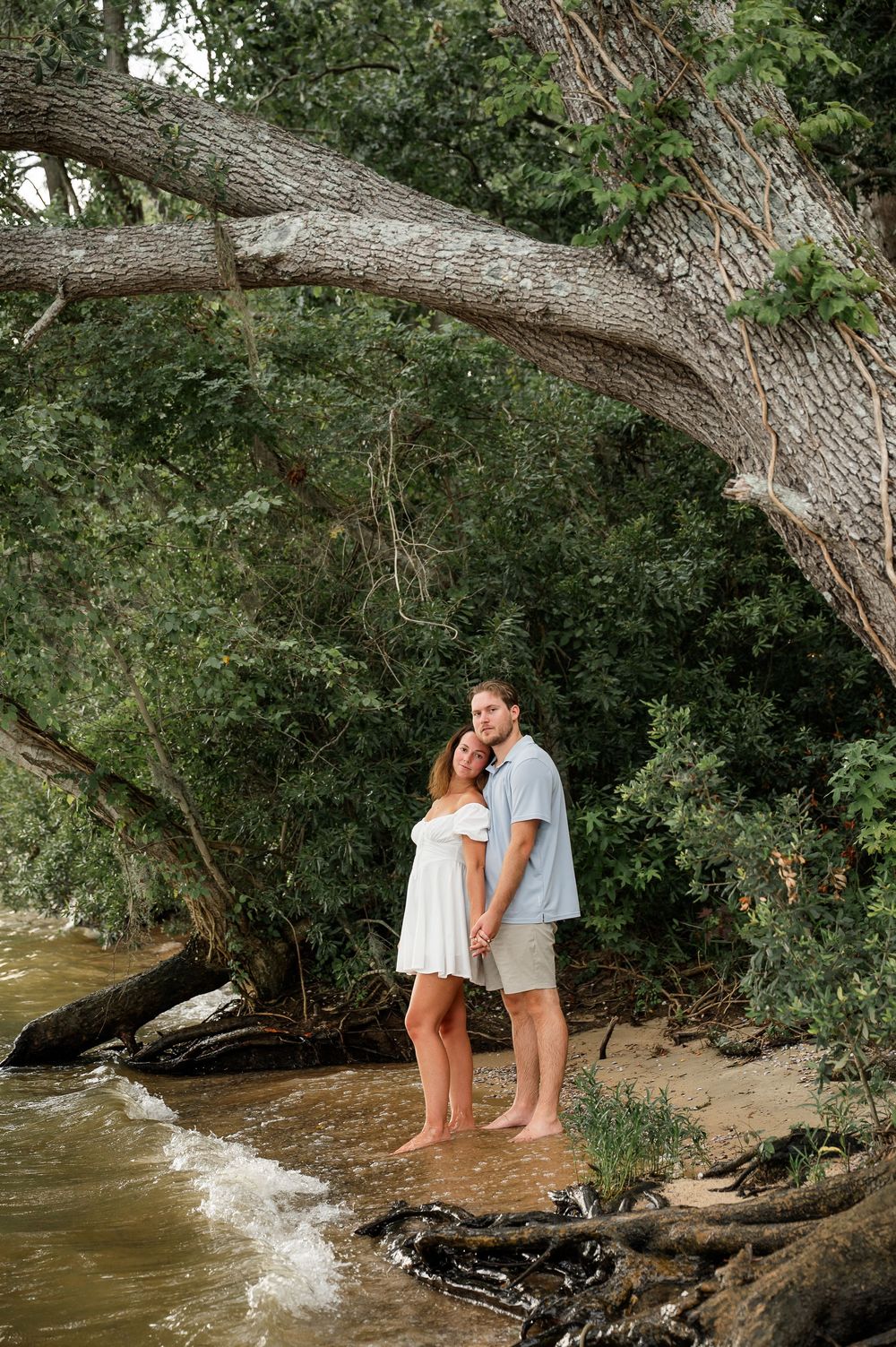 A couple stands together on a natural beach beneath an overhanging tree.