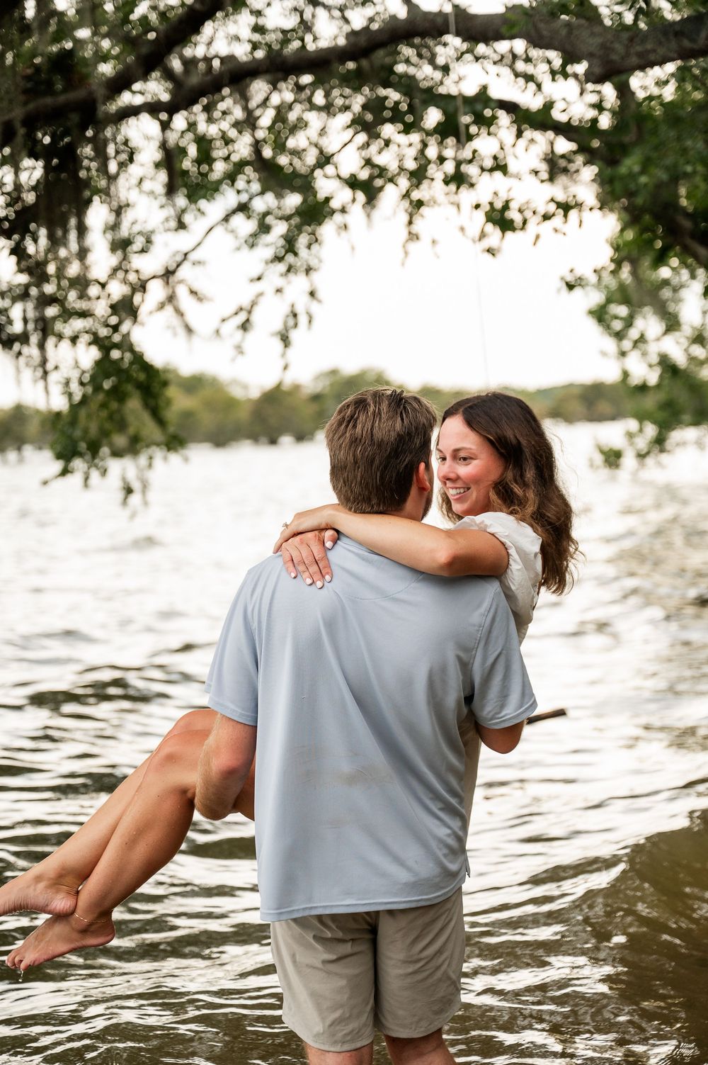 Playful couple embracing while standing in lake water with overhanging tree branches above.