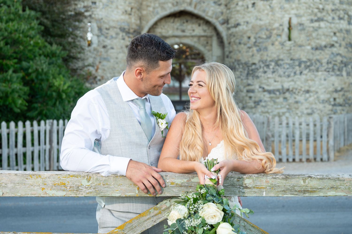 A bride and groom relax after their wedding outside Cooling Castle Barn. The evening sun lights their faces as they smile at each other.