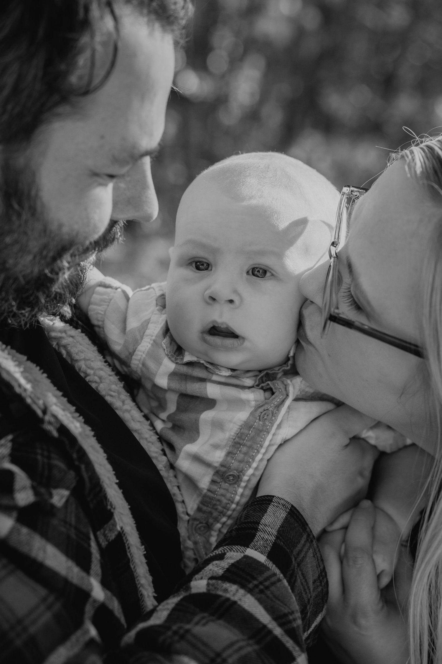 Black and white photo of bearded father and mother tenderly holding wide-eyed newborn baby outdoors.