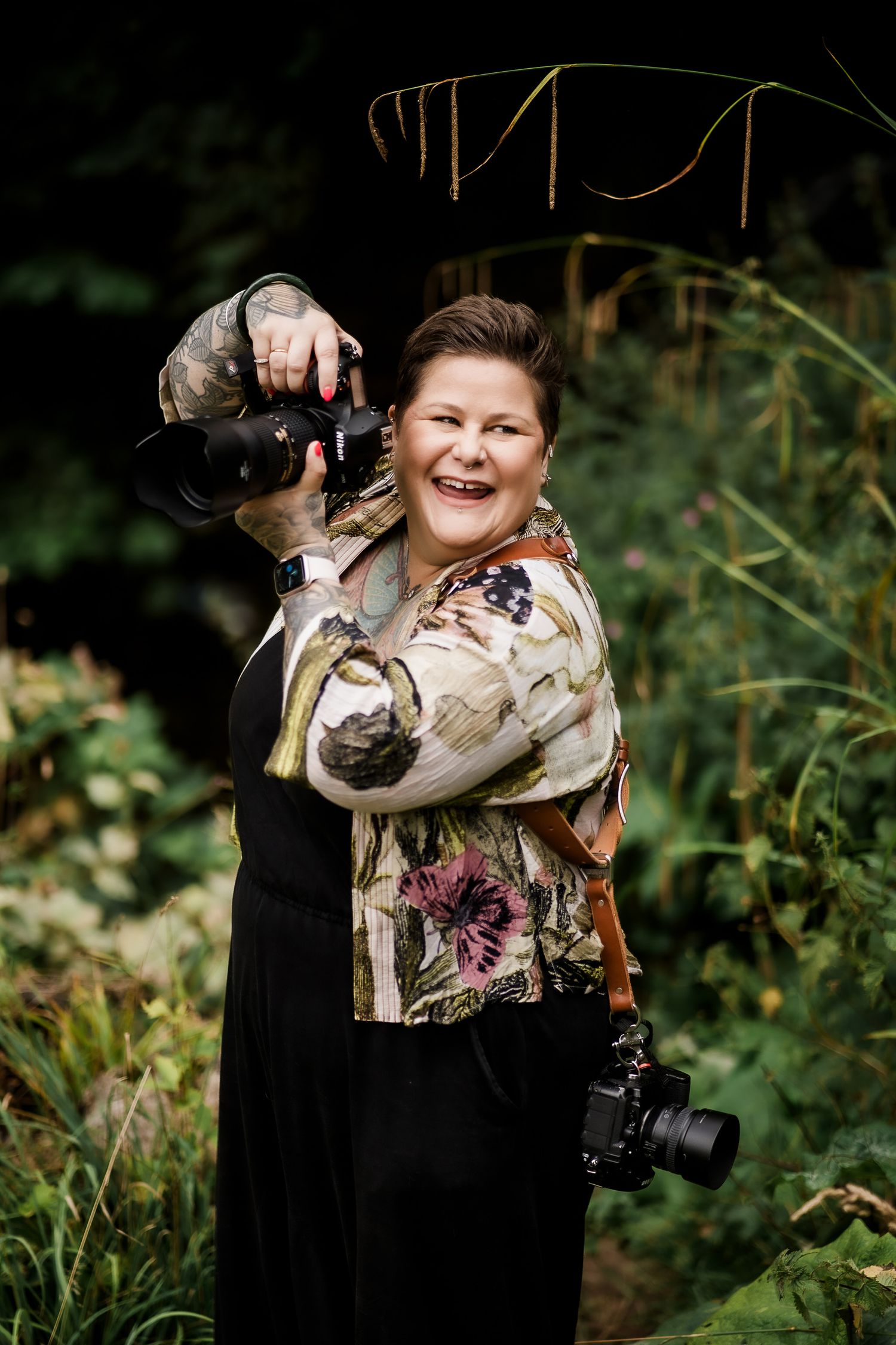 A photographer in dark clothing holds a camera while standing outdoors among green foliage.