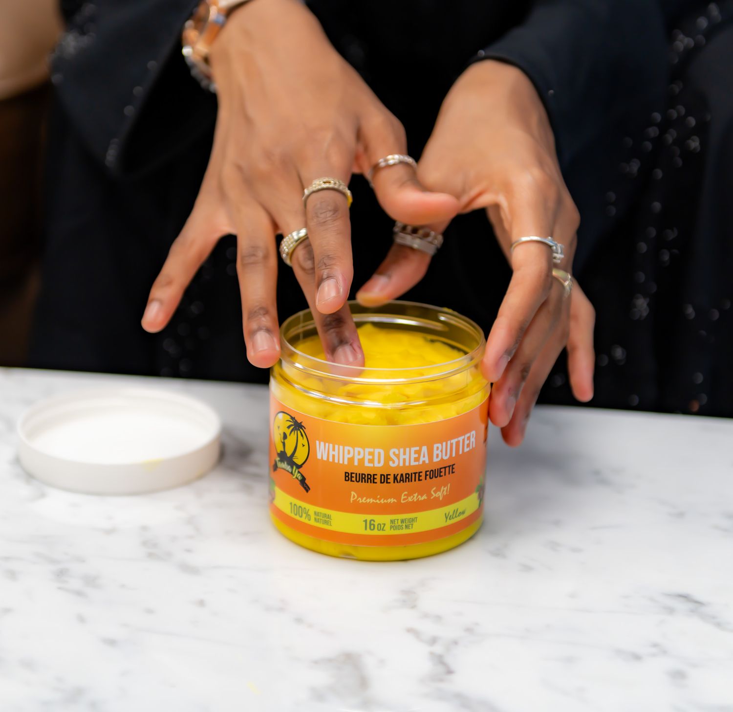Hands wearing multiple silver rings reaching for a jar of yellow whipped shea butter on a marble countertop.