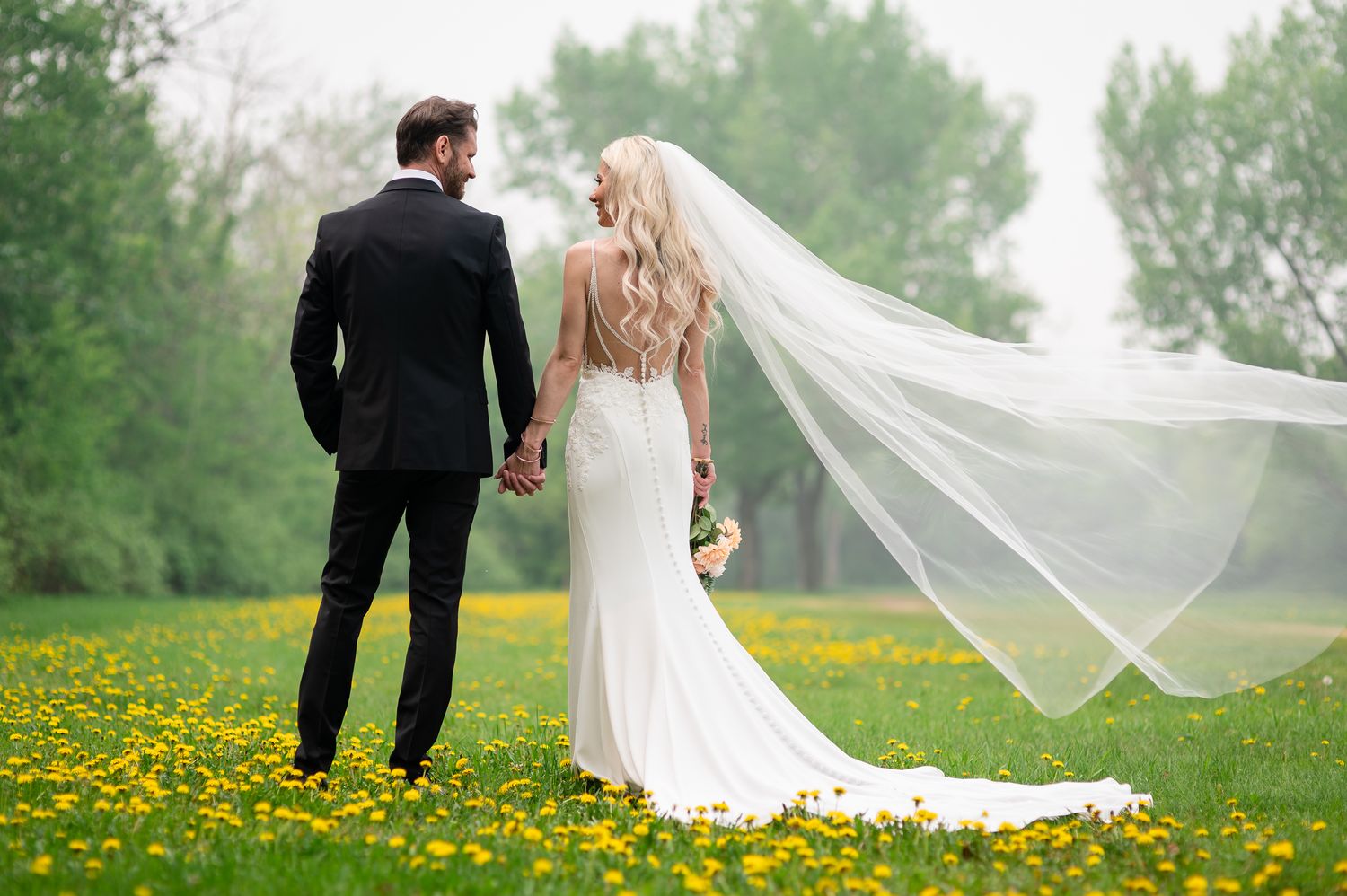 A couple in formal wedding attire shares a romantic moment together on a tree-lined path in a lush green forest setting.