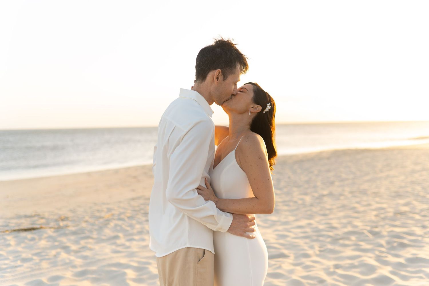 A couple in white outfits shares a romantic kiss on a sandy beach at sunset.