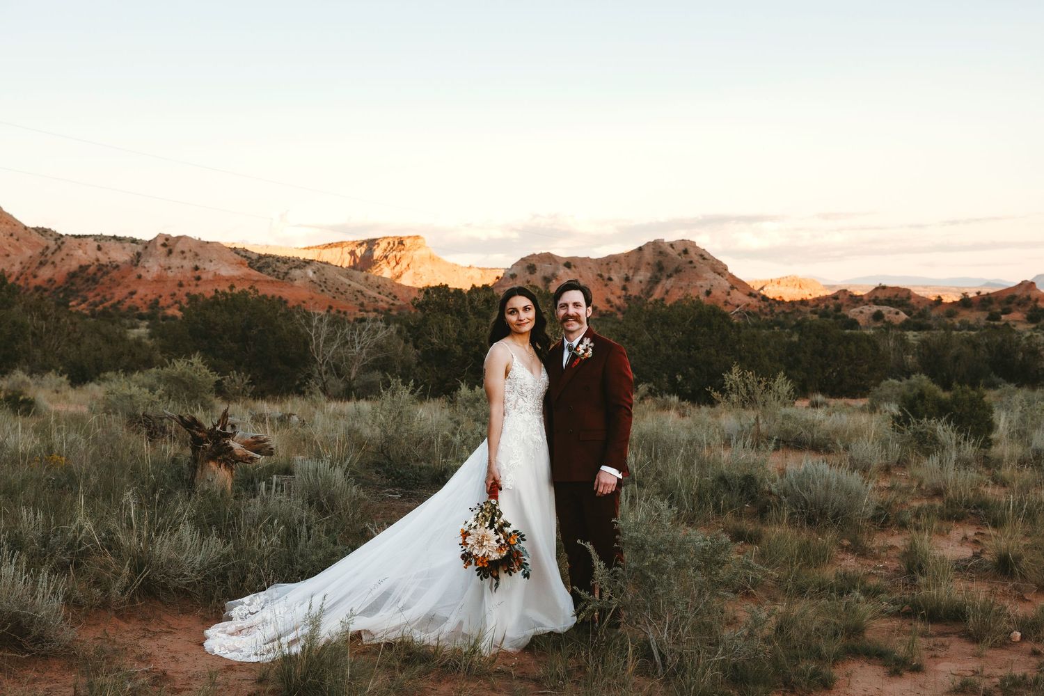 Bride and groom pose together in desert landscape with red rock formations glowing at sunset.
