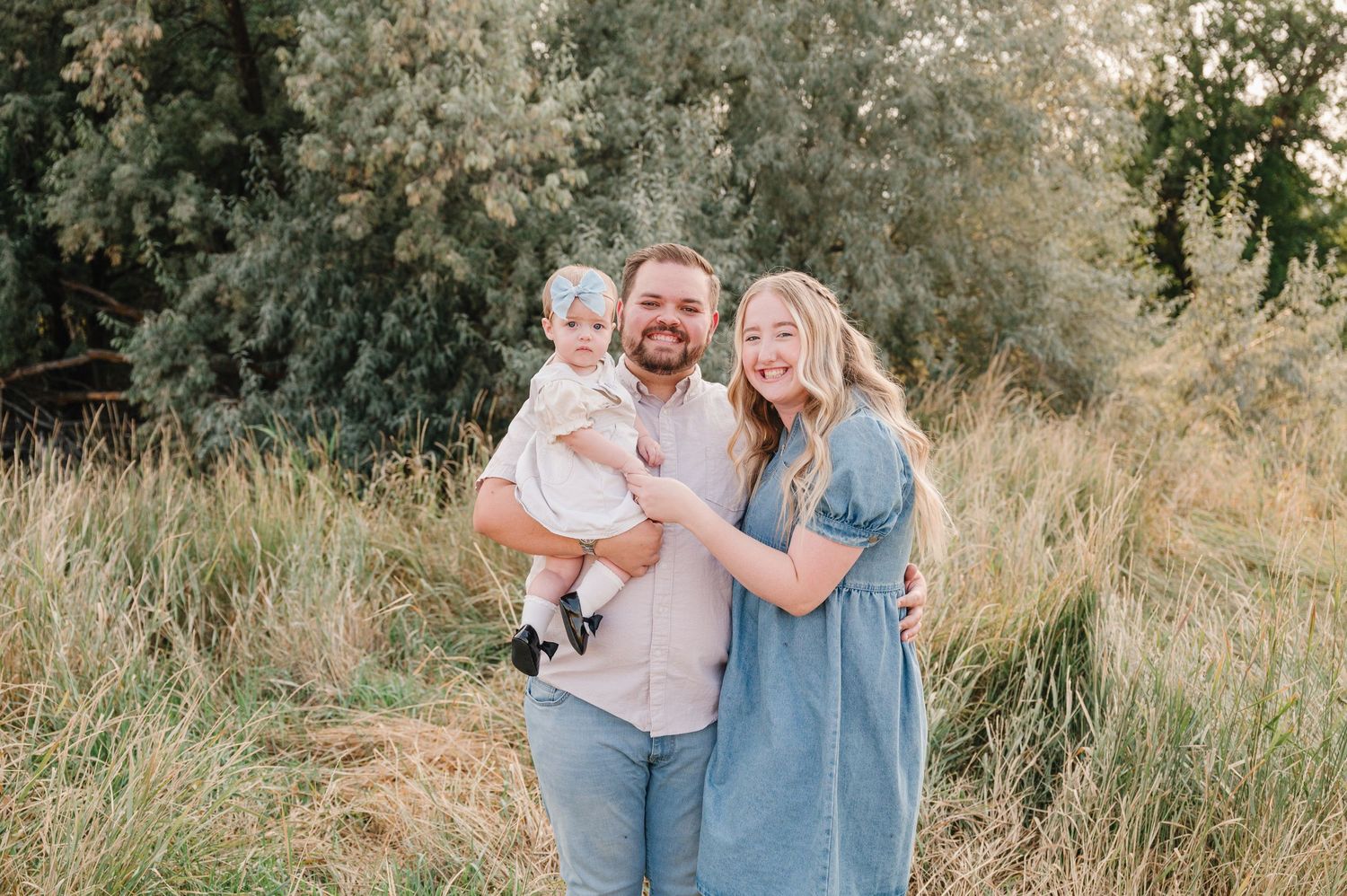 A family stands together outdoors in a field of tall grass during golden hour with soft natural lighting.
