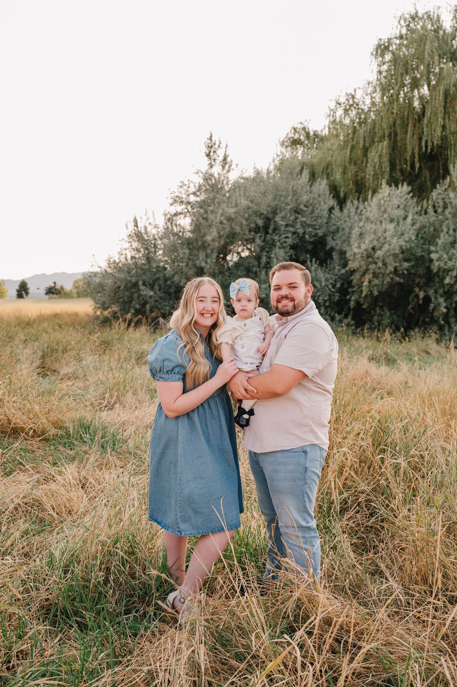 A family embraces together in a sunlit field with tall grass and trees in the background.
