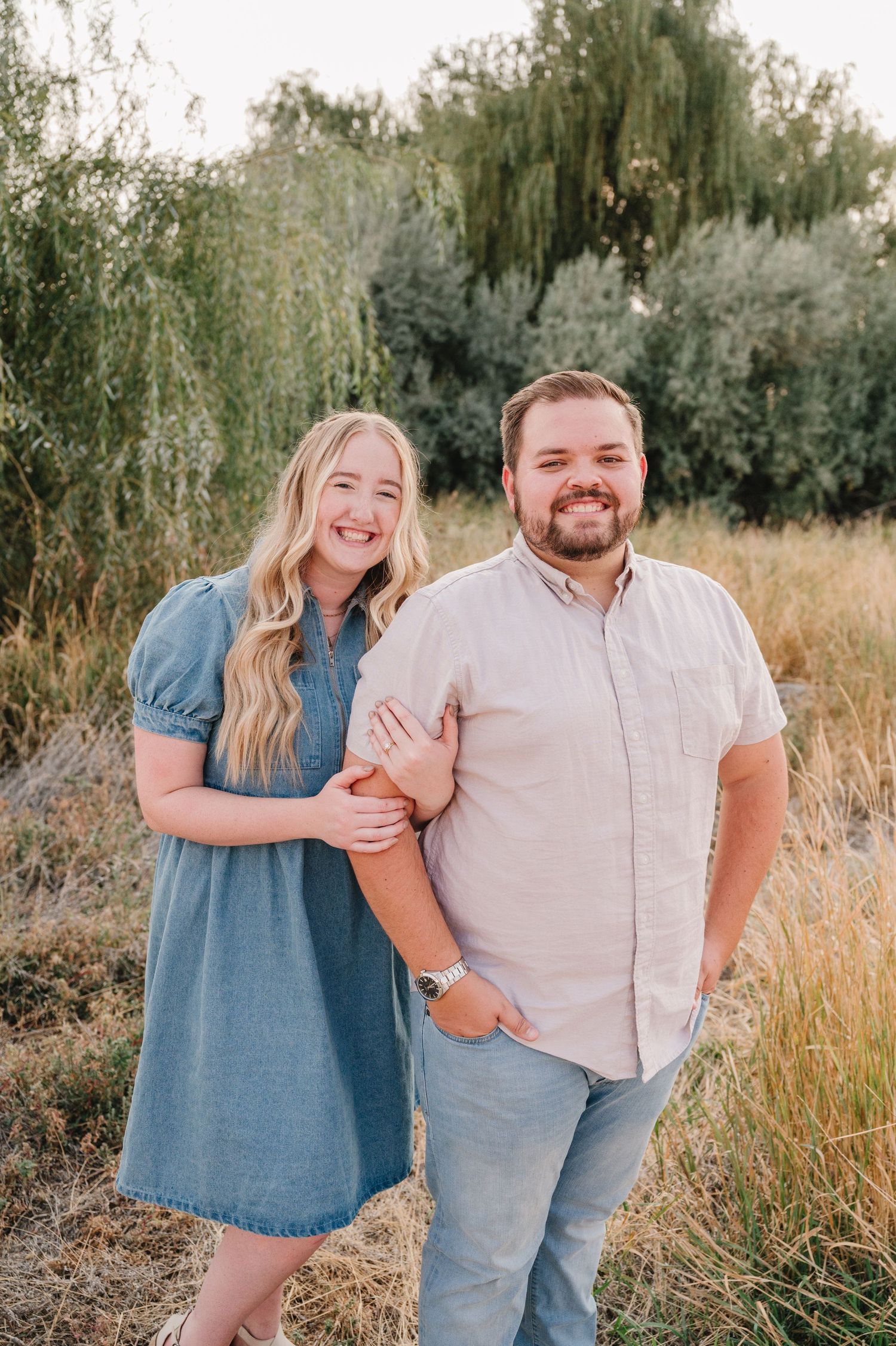 A couple in casual blue and white outfits stands together in a golden field during sunset.