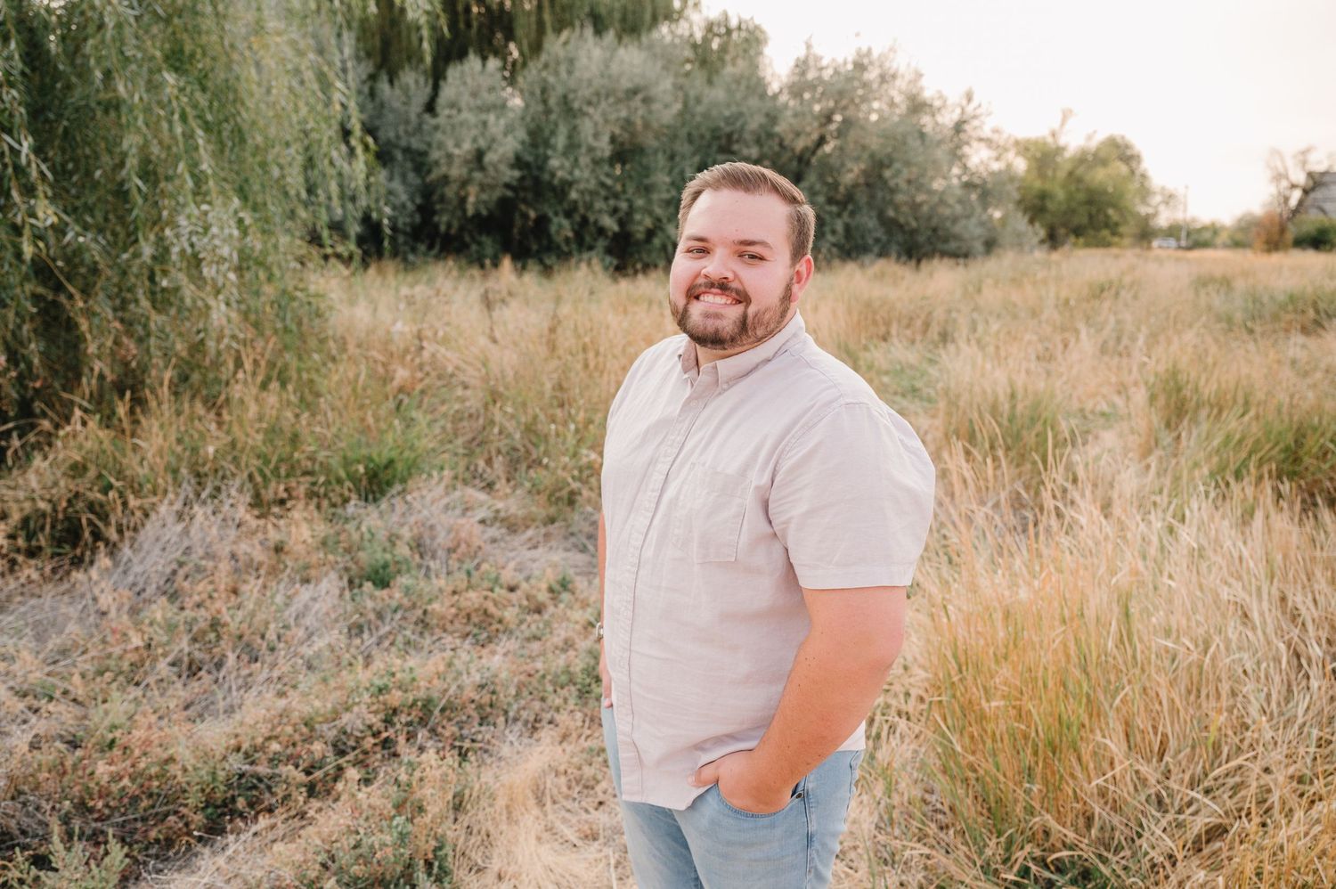 Person wearing light colored clothing poses outdoors in a field of tall golden grass at sunset.