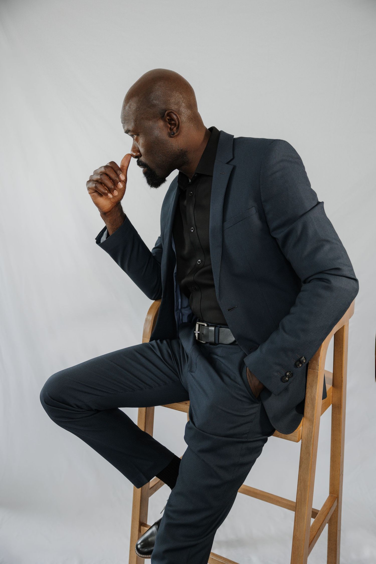 A person in a dark suit sits on a wooden stool against a white background in a contemplative pose.