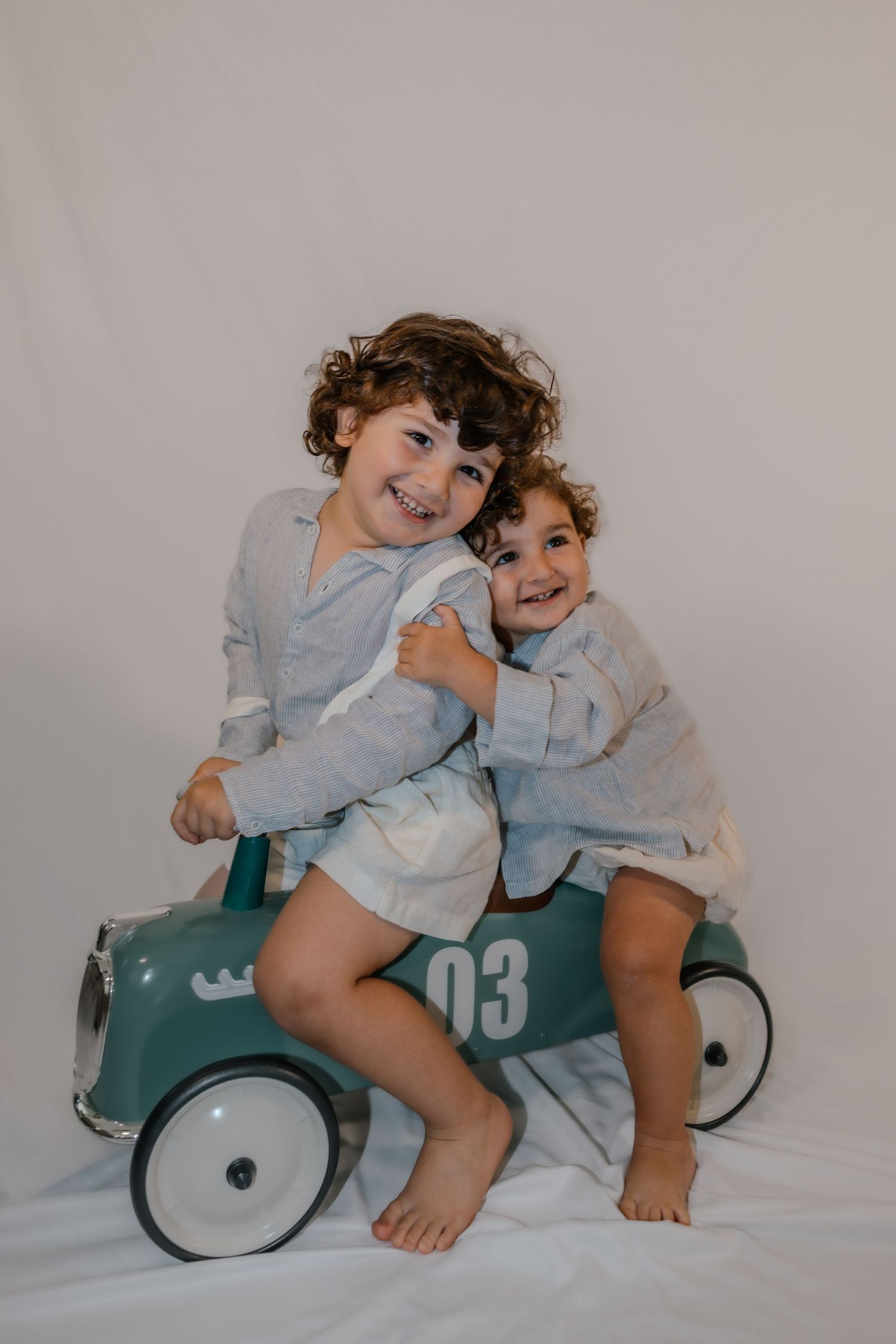 Two smiling children in matching gray outfits sitting on a teal vintage ride-on toy car numbered 03.