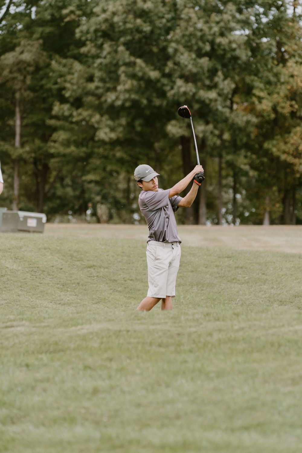A person in casual golf attire takes a swing on a grassy course with trees in the background.