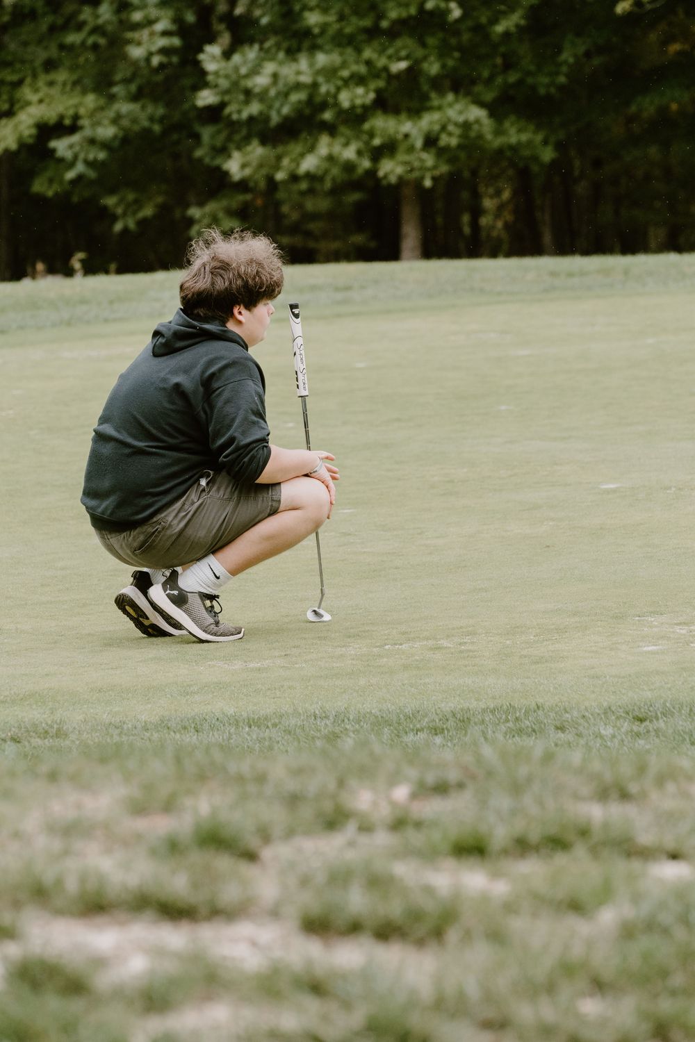 A golfer in dark clothing crouches to examine the putting surface near a flag stick.
