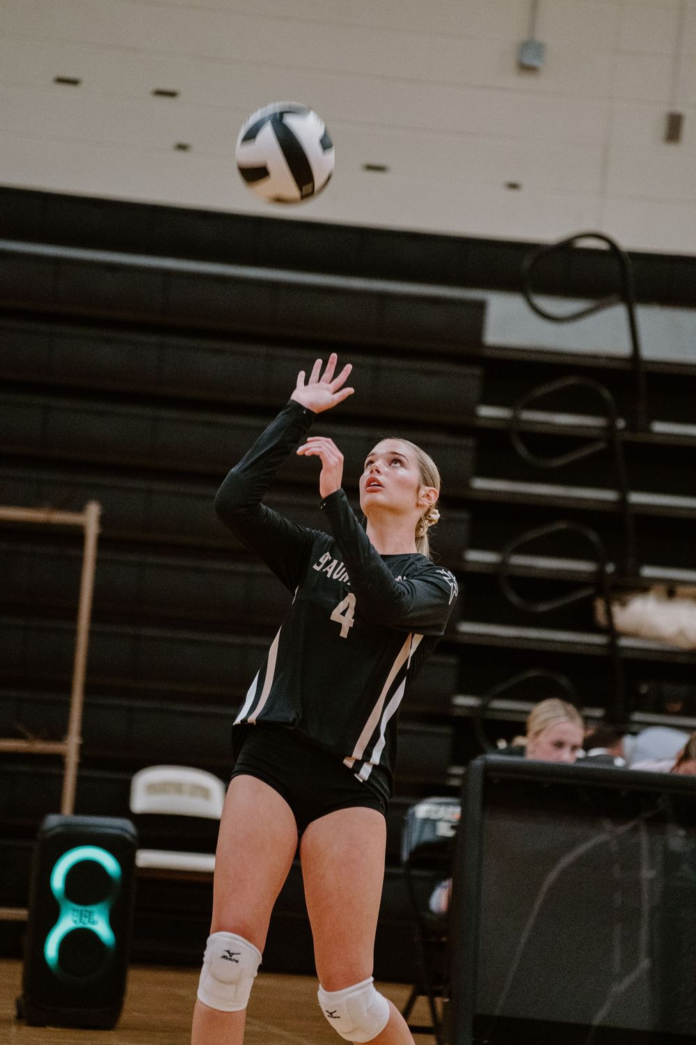 Volleyball player in black jersey number 4 jumps to serve the ball in a gymnasium.