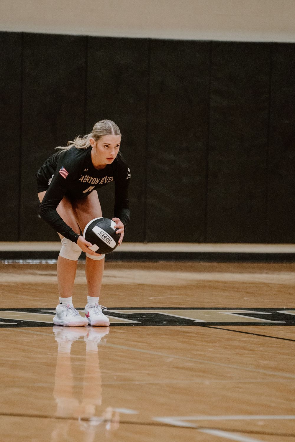Volleyball player crouches in ready position holding a volleyball on indoor court.