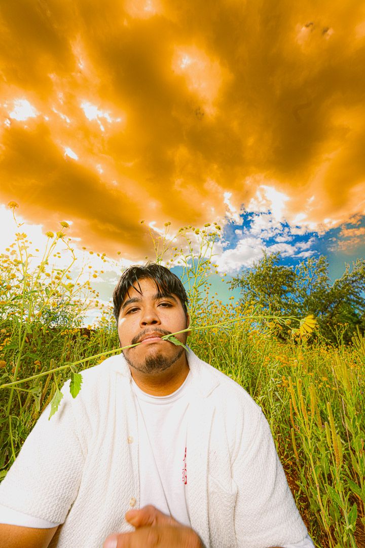 Person in white t-shirt poses playfully with grass stem in mouth against dramatic orange and blue sky in meadow.