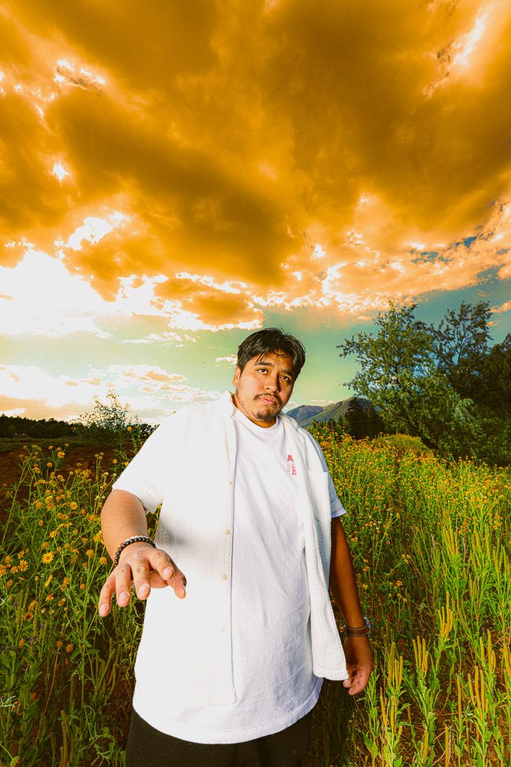 Dramatic orange sunset sky over a green field with a person in white standing among tall grass and wildflowers.