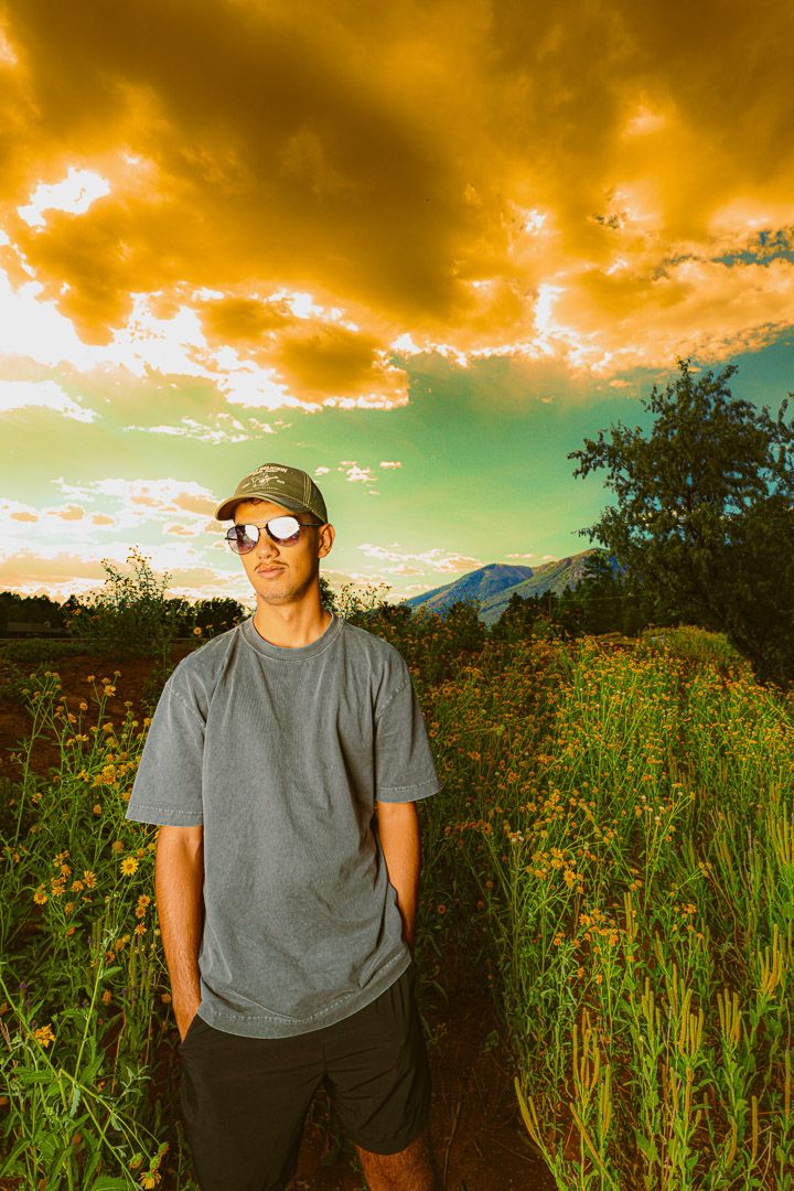 Dramatic orange sunset sky over a green field with someone in a gray t-shirt standing among tall grass and wildflowers.
