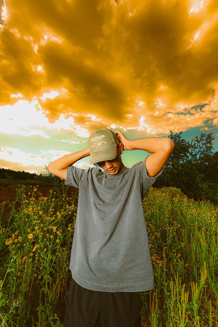 Person in gray t-shirt stands in wildflower field at sunset with dramatic orange clouds overhead.