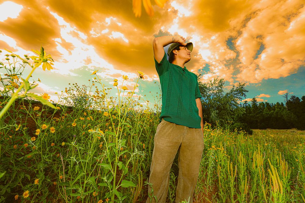 Silhouetted figure stands in tall grass field against dramatic orange and yellow sunset sky.