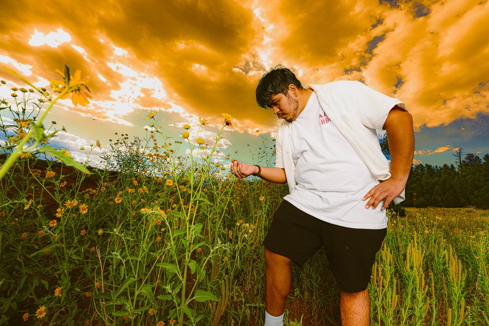 A person stands in a lush green field under dramatic orange and yellow clouds at sunset.