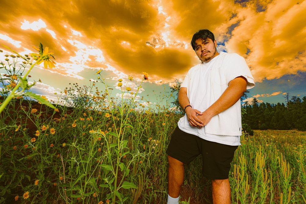 A person in white clothing stands in a lush green field against a dramatic orange and cloudy sunset sky.