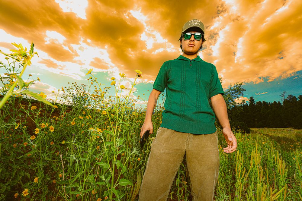 A person in a green shirt stands in a tall grass field against a dramatic orange and blue sunset sky.