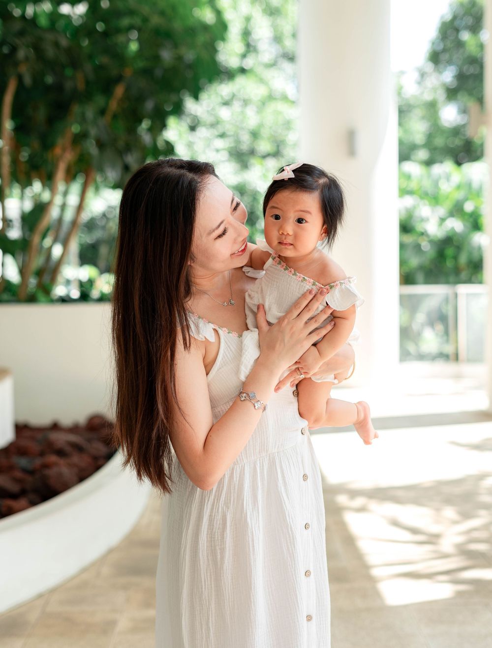 A parent in white lovingly gazes at their baby while standing in front of lush greenery on a bright porch.