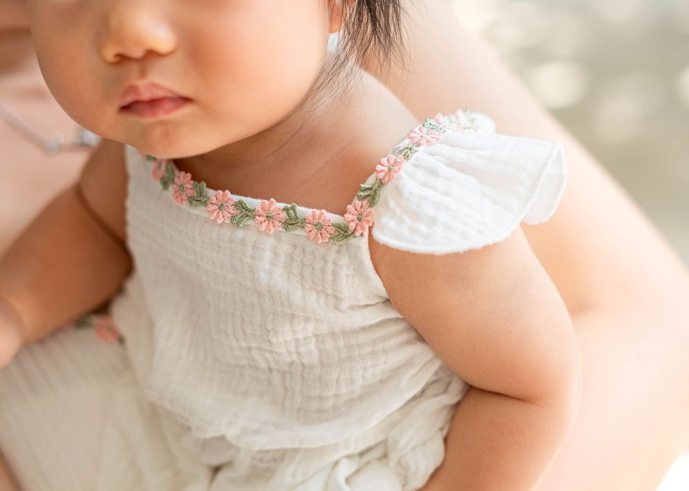 A baby in a white dress sits on a soft surface while looking away from the camera.
