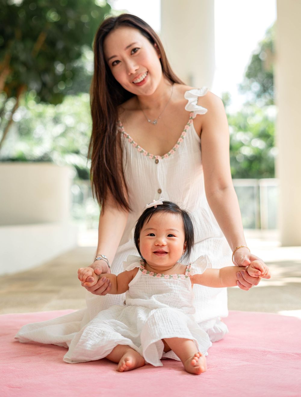 A woman in white dress sits with a baby on a pink blanket in a bright indoor setting with natural light.