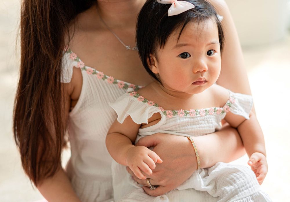 A baby wearing a white dress sits on a soft white surface in a bright, airy setting.
