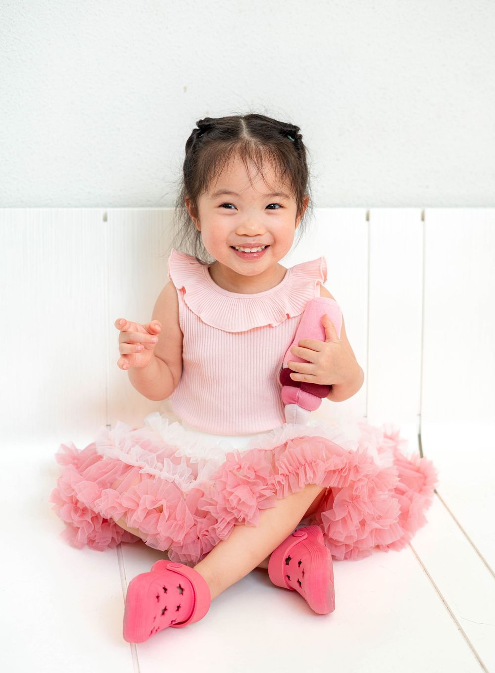 A child in a pink tutu dress sits on a white wooden floor with a joyful expression.
