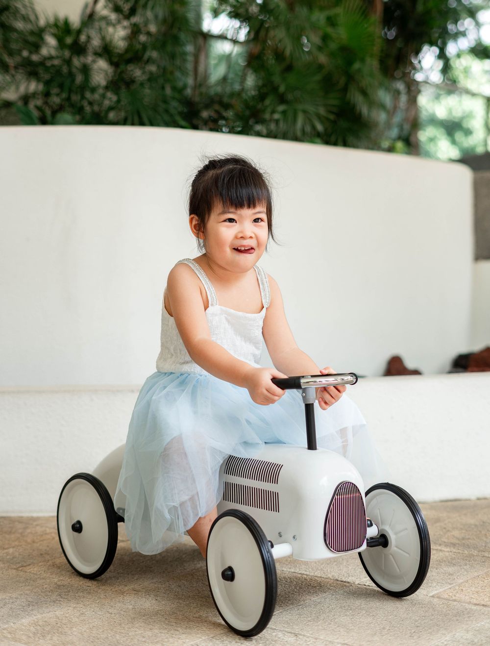 A child in a white dress rides a minimalist wooden balance bike on a patio in a sequence of playful shots.