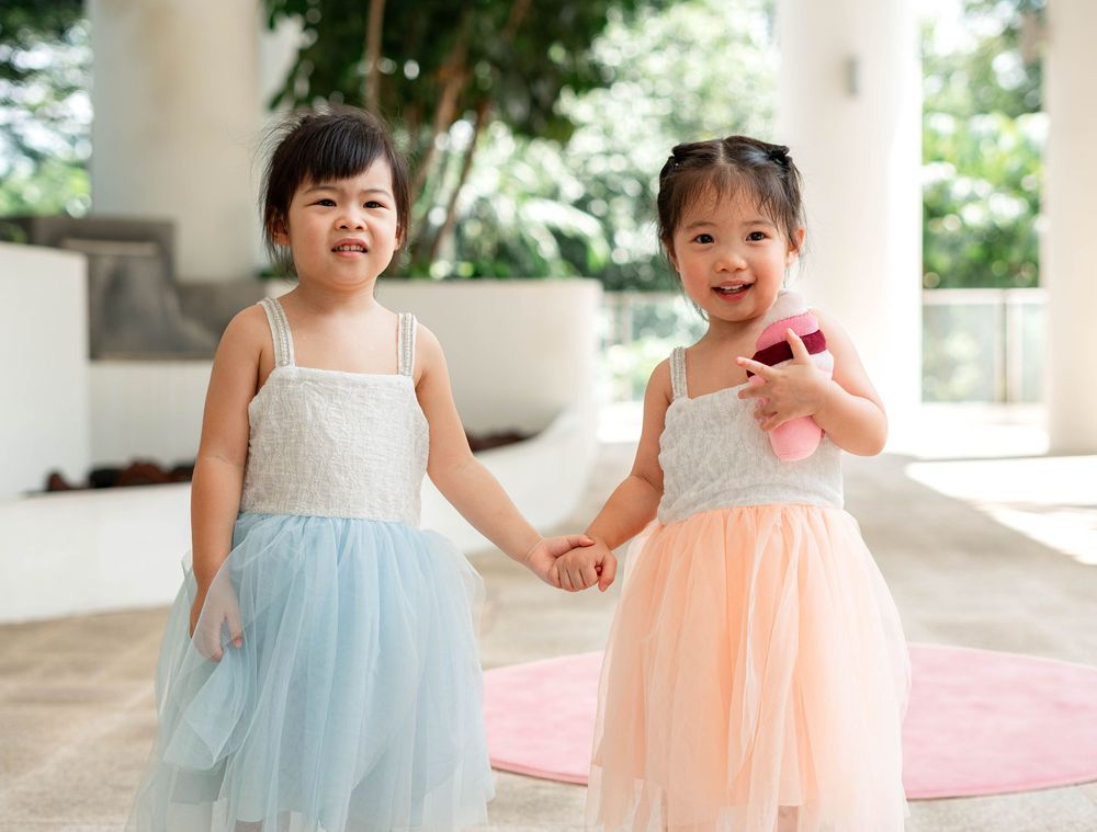Two children in pastel blue and peach dresses play together in a bright indoor space.