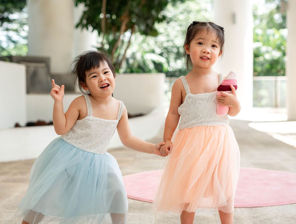 Two toddlers in tulle dresses laugh and play together in a bright indoor space.