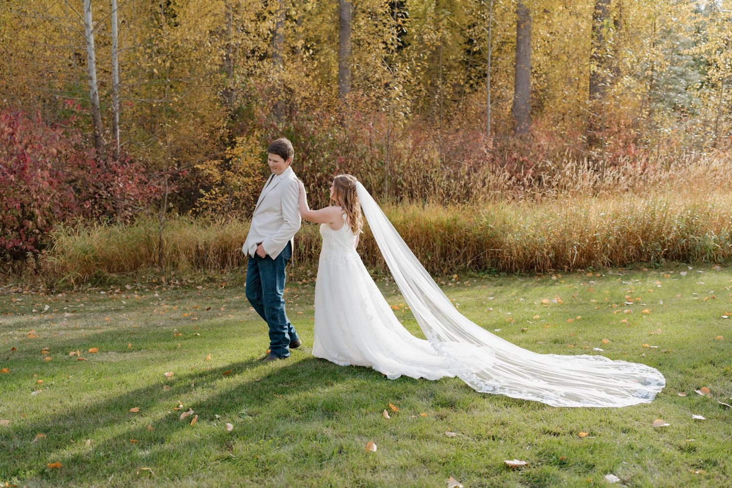A couple poses in an autumn landscape with golden trees and red foliage while wearing wedding attire on a grassy lawn.