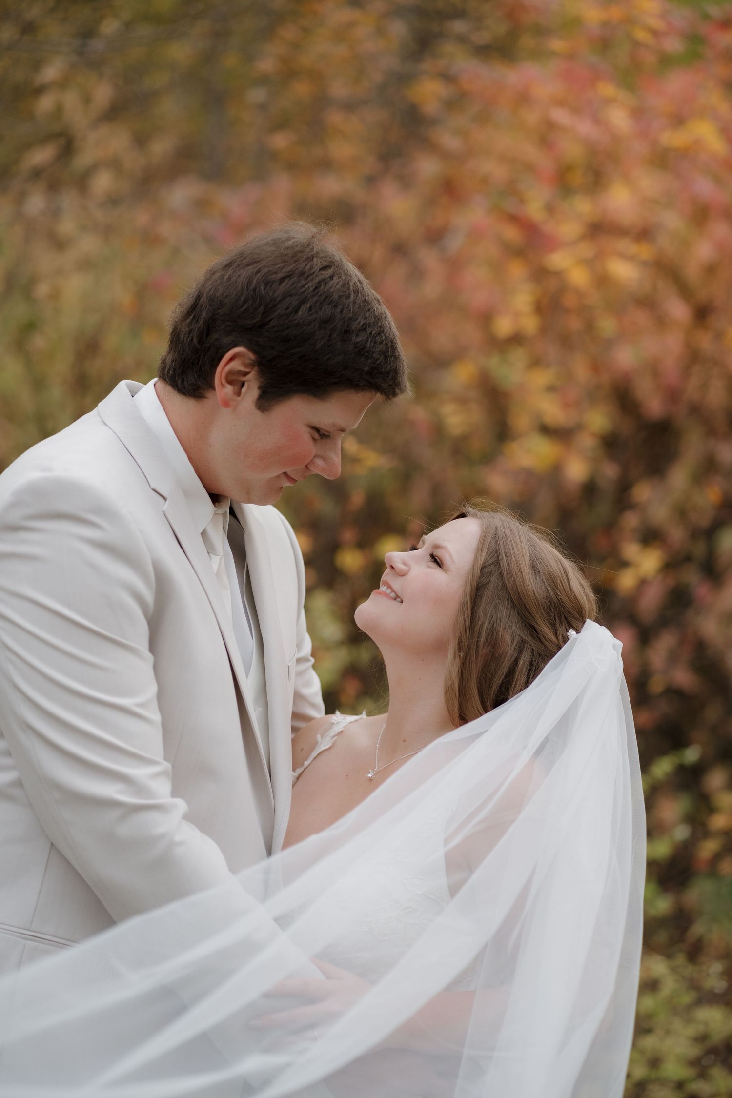 A tender romantic wedding moment with a bride in flowing veil and groom in light suit against autumn foliage.