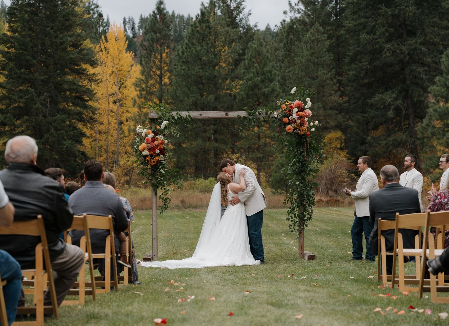 An intimate autumn outdoor wedding ceremony takes place under a rustic wooden arch adorned with floral arrangements.