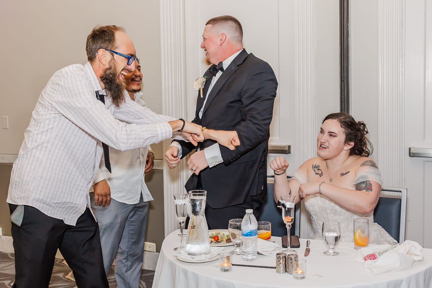 A candid moment at a wedding reception shows guests interacting at a decorated table with drinks and place settings.