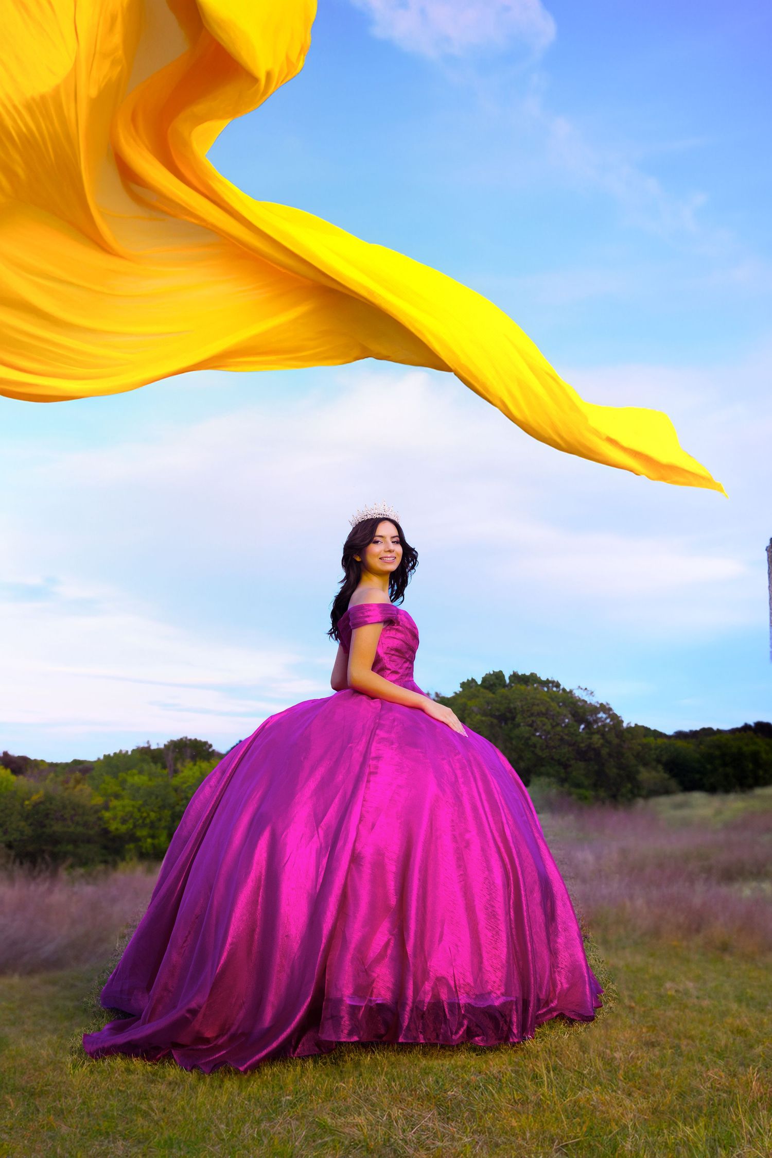 A person in a flowing magenta ballgown poses dramatically in a field at sunset with yellow fabric billowing overhead.