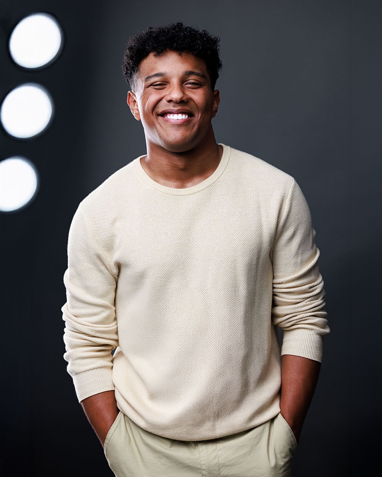Person wearing cream colored sweater smiling against dark background in studio lighting setup.