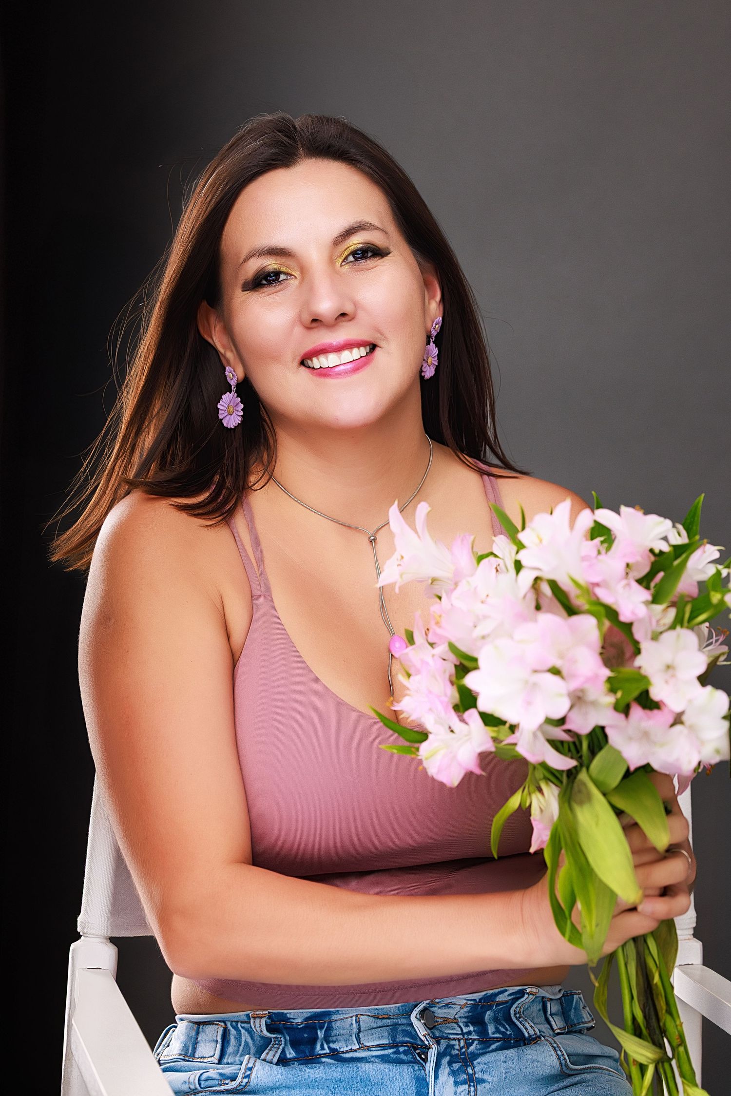 A person in a pink tank top and jeans holds a bouquet of pink flowers while posing against a dark background.